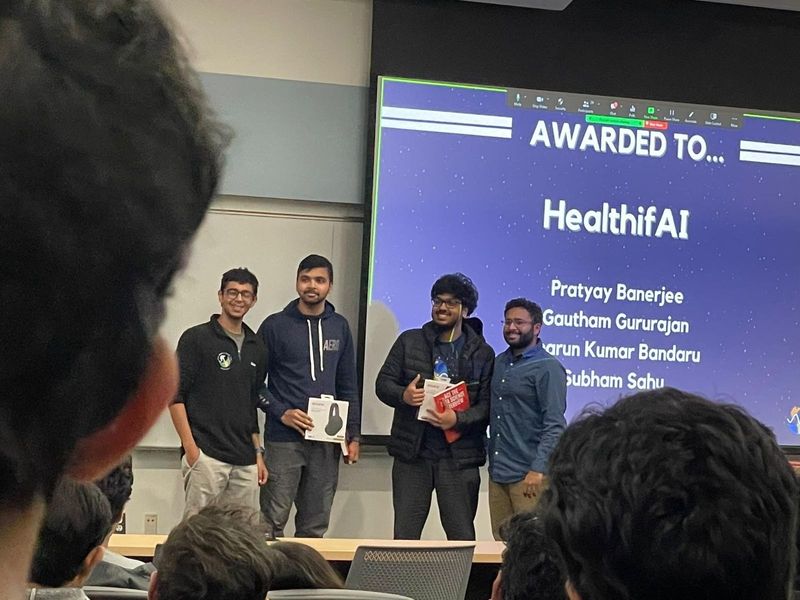 On stage at a hackathon prize announcement, with the slide reading Awarded to HealthifAI above the team's names